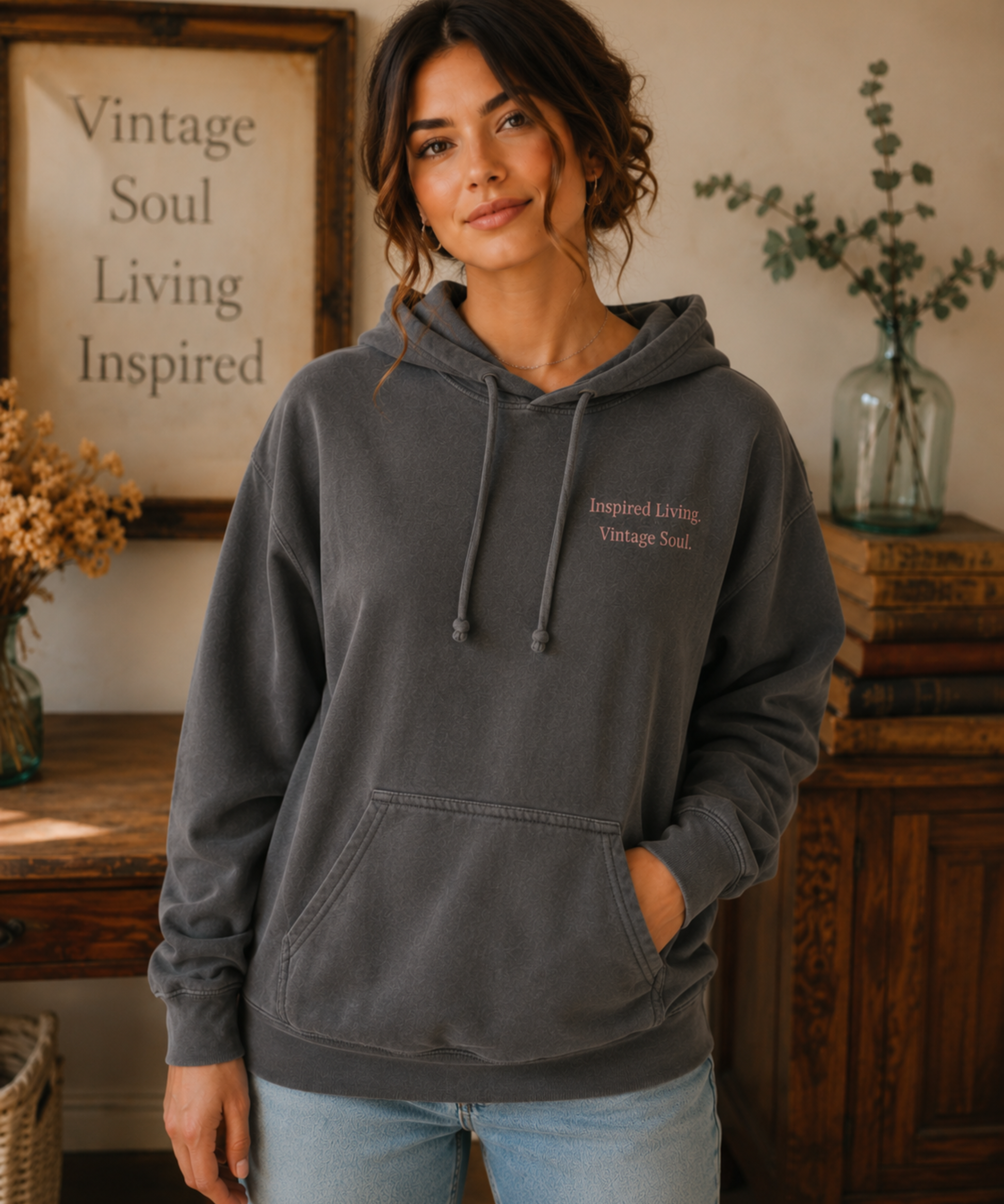 Woman wearing a Comfort Colors Pepper hoodie featuring the small "Inspired Living. Vintage Soul." left chest design in a warm vintage-inspired home with rustic wood furniture and soft neutral décor.