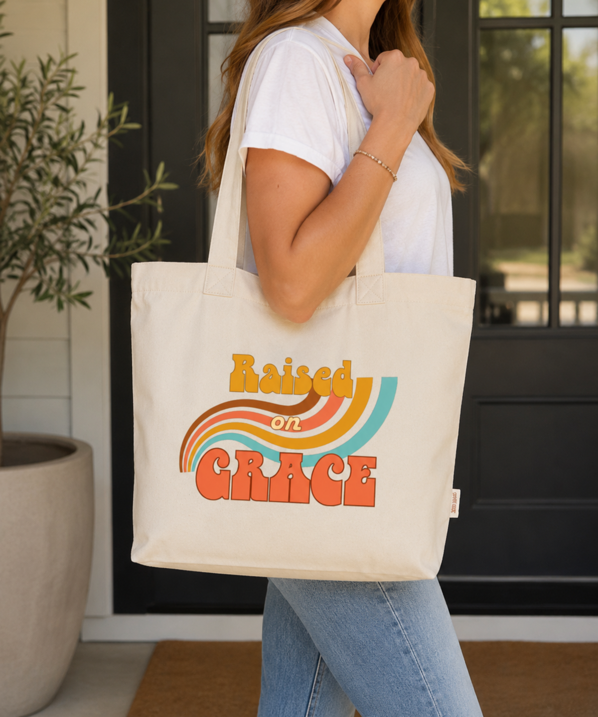 Woman carrying an Ivory Comfort Colors Everyday Zip Tote featuring the retro Raised on Grace design in warm mustard, coral, brown, cream, and aqua colors. Casual lifestyle mockup showcasing a vintage-inspired faith tote with a nostalgic rainbow graphic and uplifting Christian message.