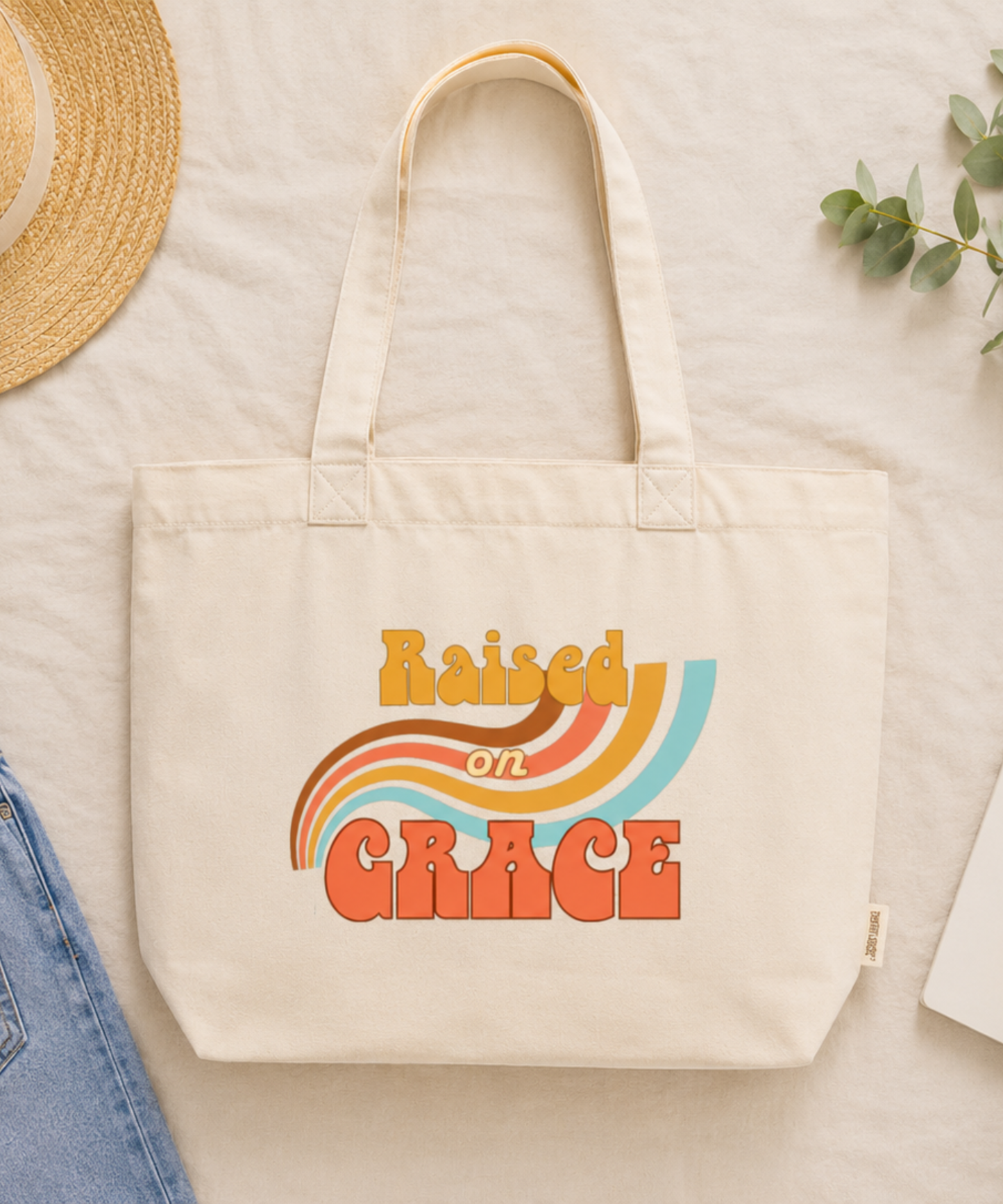 Ivory Comfort Colors Everyday Zip Tote displayed in a cozy flat lay with denim jeans, straw hat, greenery, notebook, and coffee. Features the retro Raised on Grace design in vintage-inspired mustard, coral, brown, cream, and aqua tones, creating a warm nostalgic faith-inspired aesthetic.