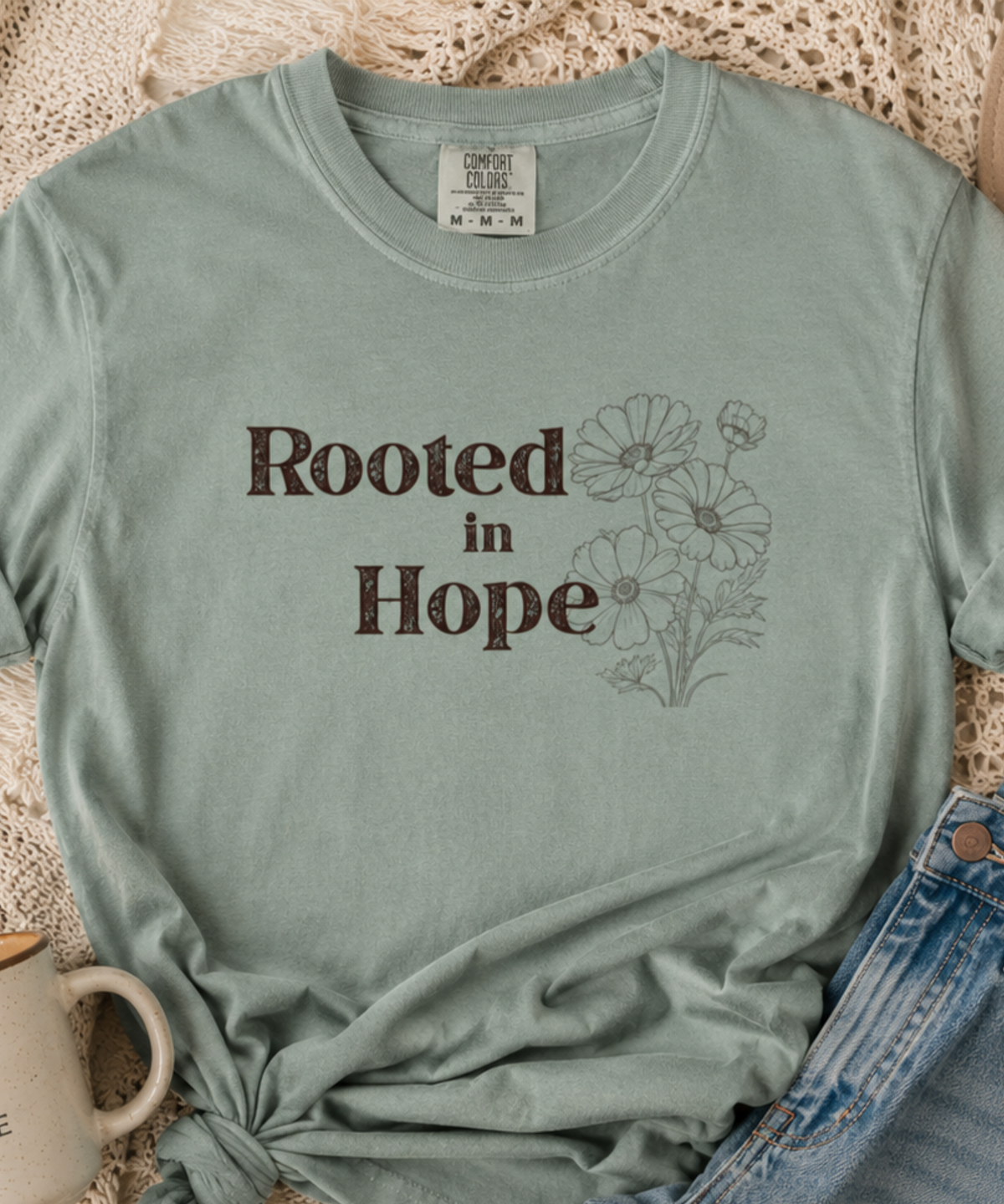 Vintage-inspired flat lay featuring a bay green “Rooted in Hope” graphic tee styled with denim jeans, neutral textures, eucalyptus stems, and cozy soulful accessories.