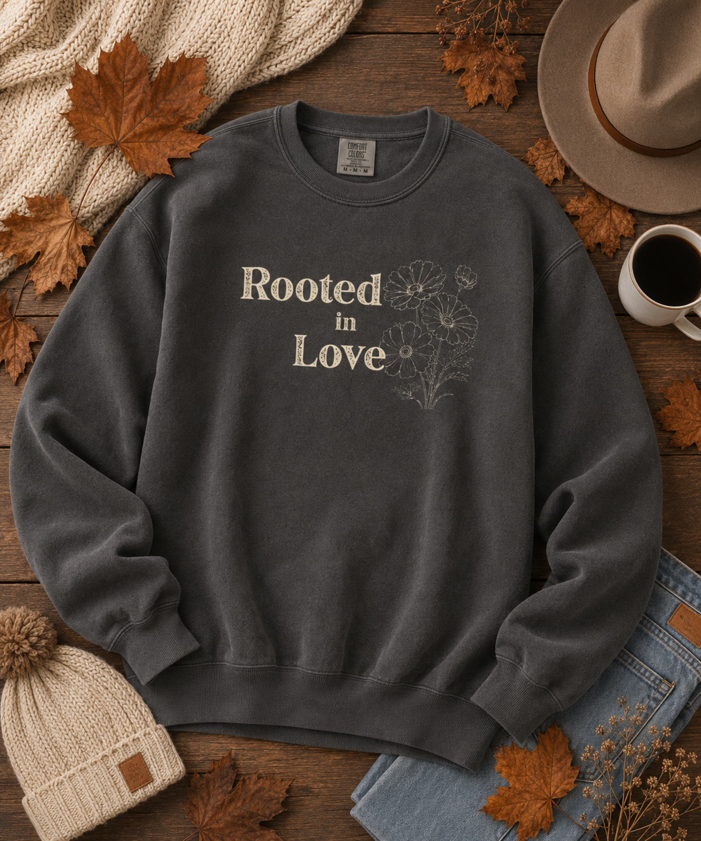 A pepper comfort colors crewneck sweatshirt with 'Rooted in Love' and a floral line drawing printed on the chest, styled in a flat lay with autumn leaves, a hat, a beanie, jeans, a mug of coffee, and a knit scarf on a wooden surface.