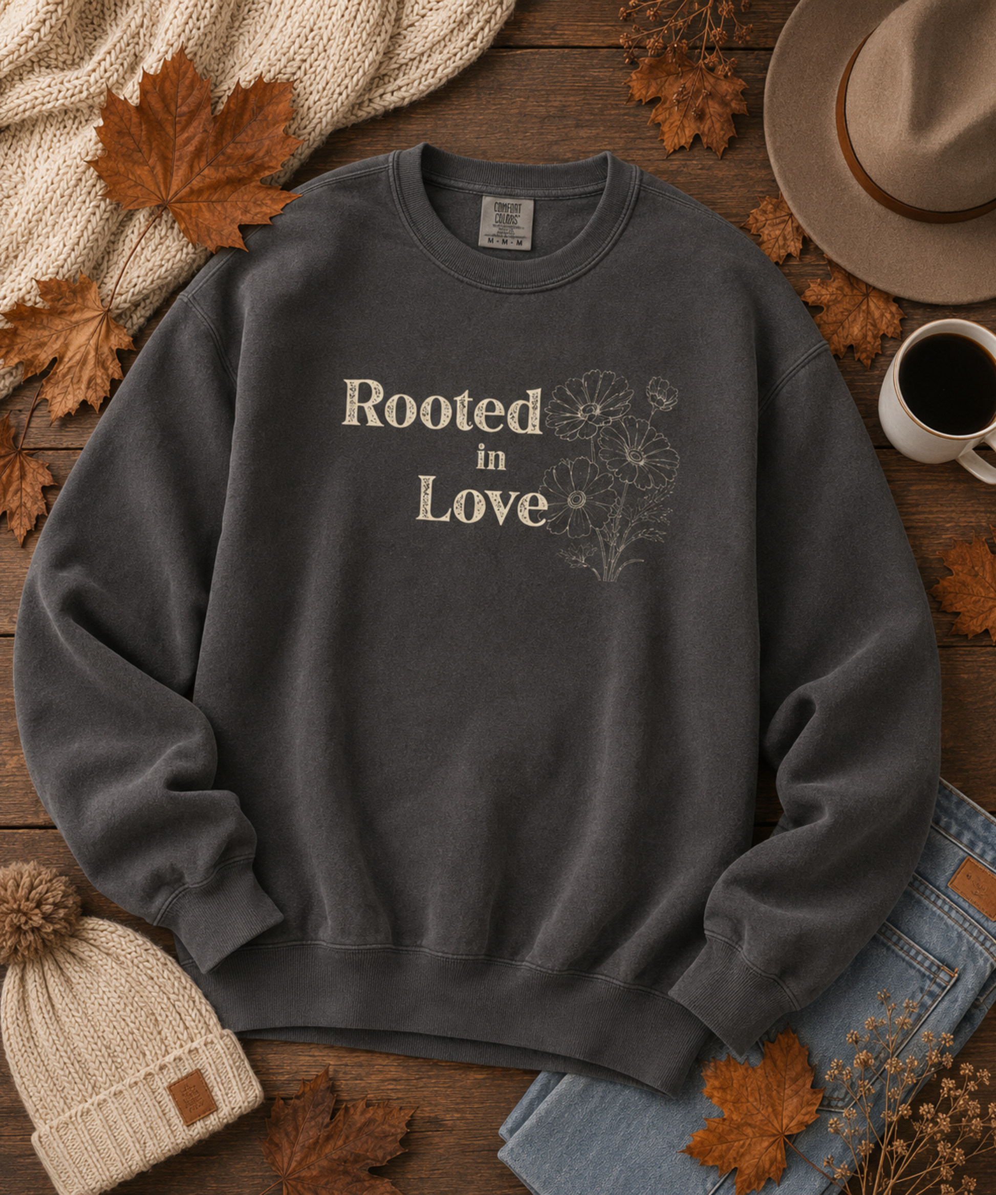 A pepper comfort colors crewneck sweatshirt with 'Rooted in Love' and a floral line drawing printed on the chest, styled in a flat lay with autumn leaves, a hat, a beanie, jeans, a mug of coffee, and a knit scarf on a wooden surface.