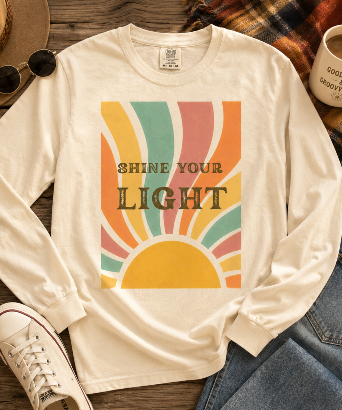 An ivory comfort colors long-sleeve t-shirt with a retro sunburst graphic and the text 'SHINE YOUR LIGHT' printed on the front. The shirt is styled in a flat-lay arrangement on a wooden surface with a plaid scarf, sunglasses, a hat, a coffee mug, and blue jeans.