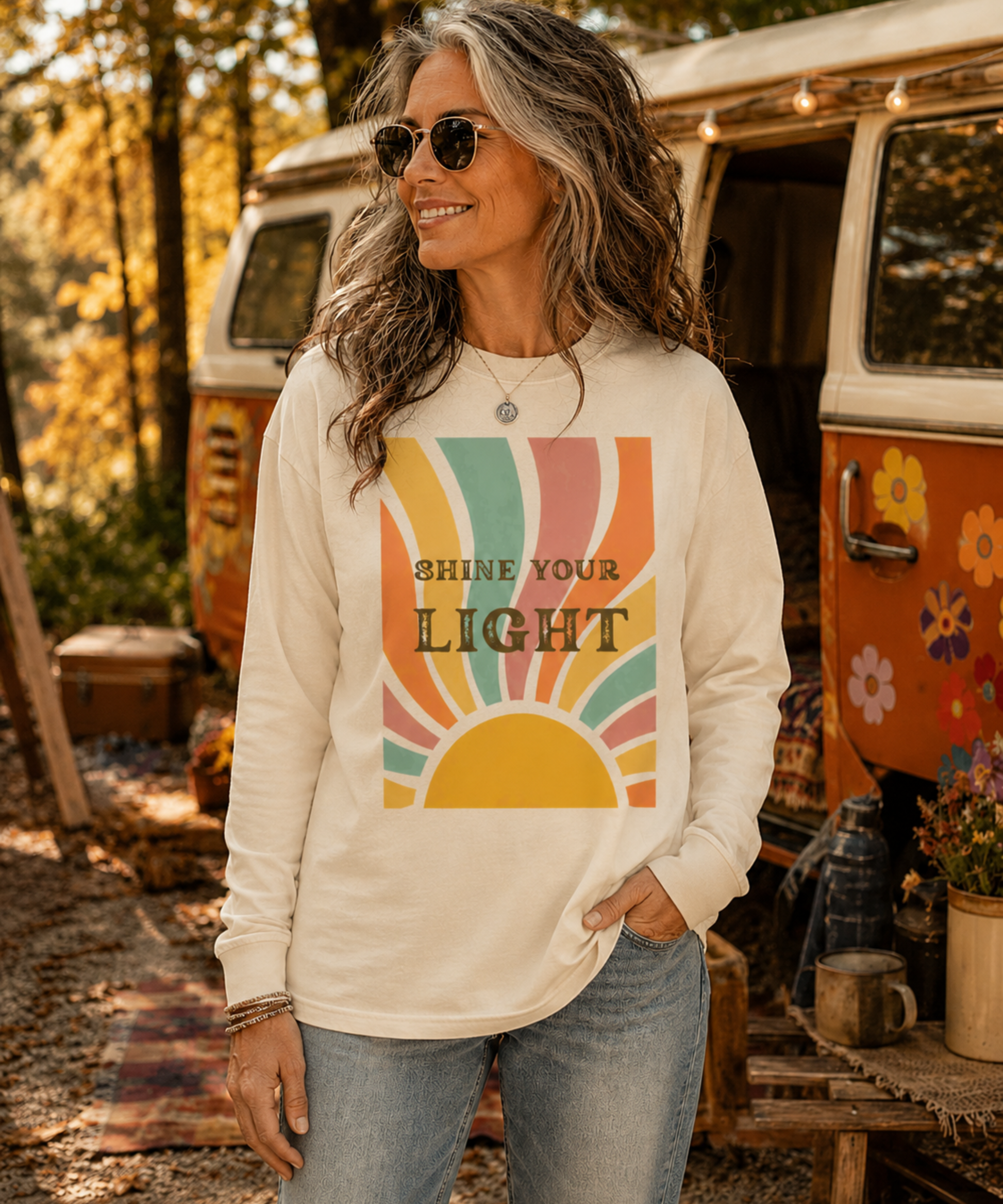 A woman wearing an ivory comfort colores long-sleeve t-shirt with a retro multi-colored sun graphic and the text 'SHINE YOUR LIGHT' in front of a vintage orange and white camper van in an outdoor campsite setting.