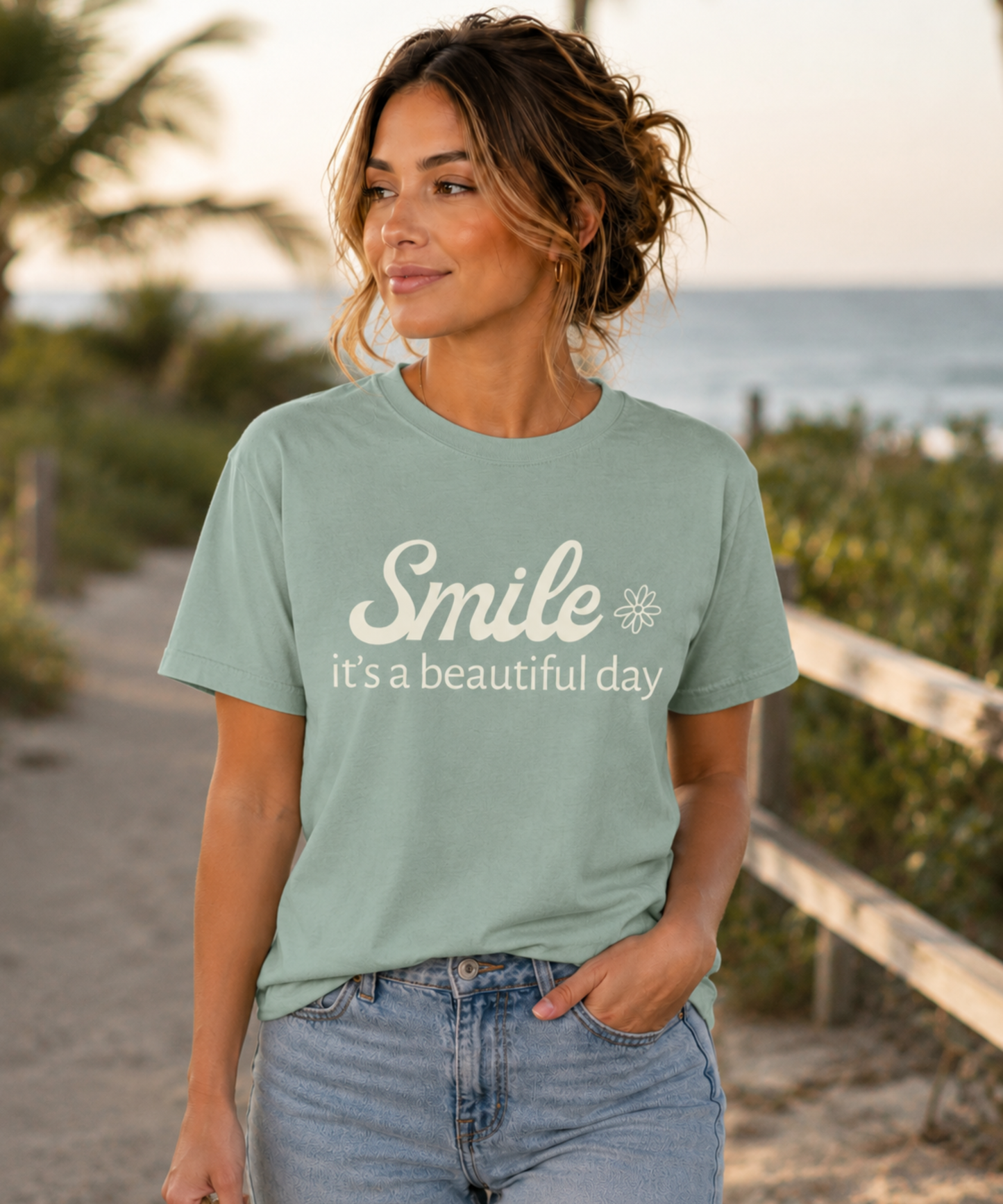 A woman wearing a bay comfort colors graphic t-shirt with the text 'Smile it's a beautiful day' in cream, standing on a beach path with the ocean and palm trees in the background.