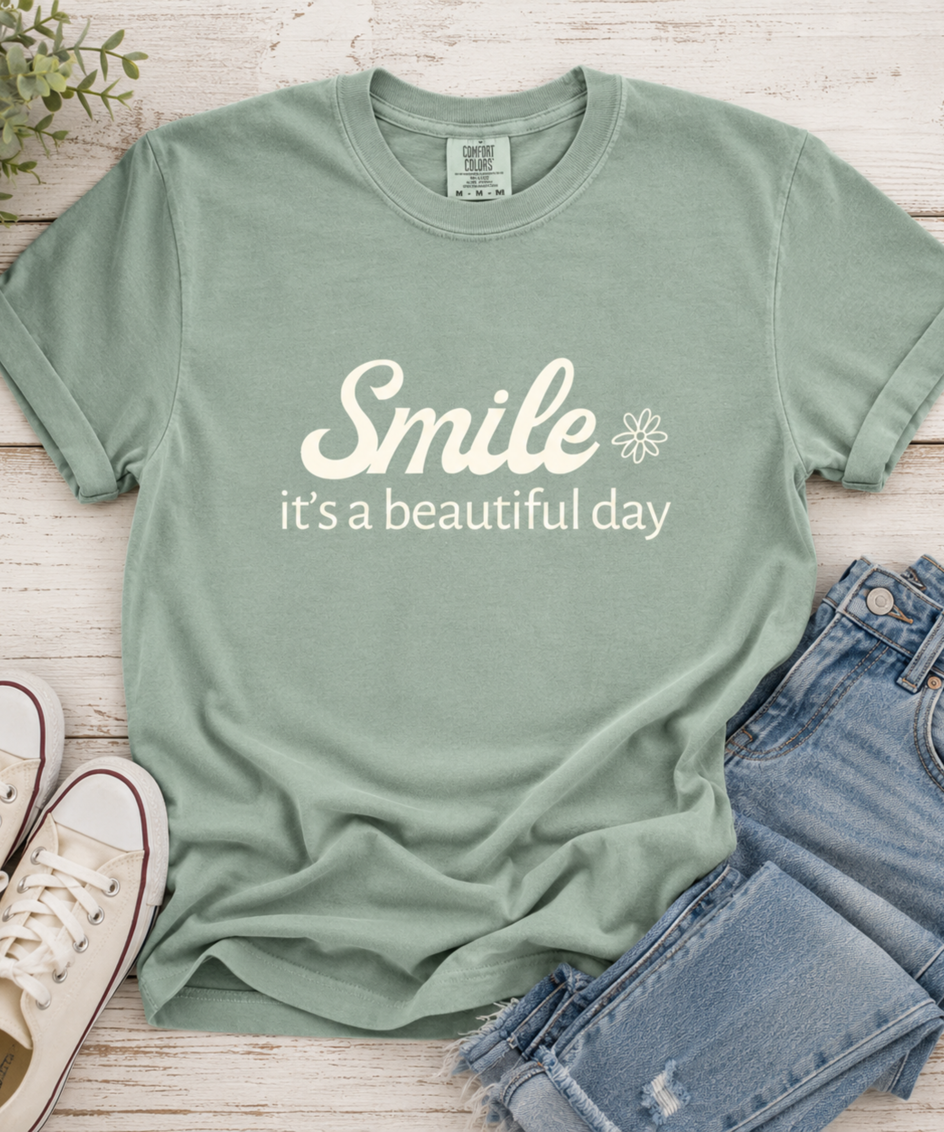 A Bay comfort colors graphic t-shirt with the text 'Smile it's a beautiful day' in cream, styled in a flat lay with blue jeans, white sneakers, and a small plant on a white wood background.