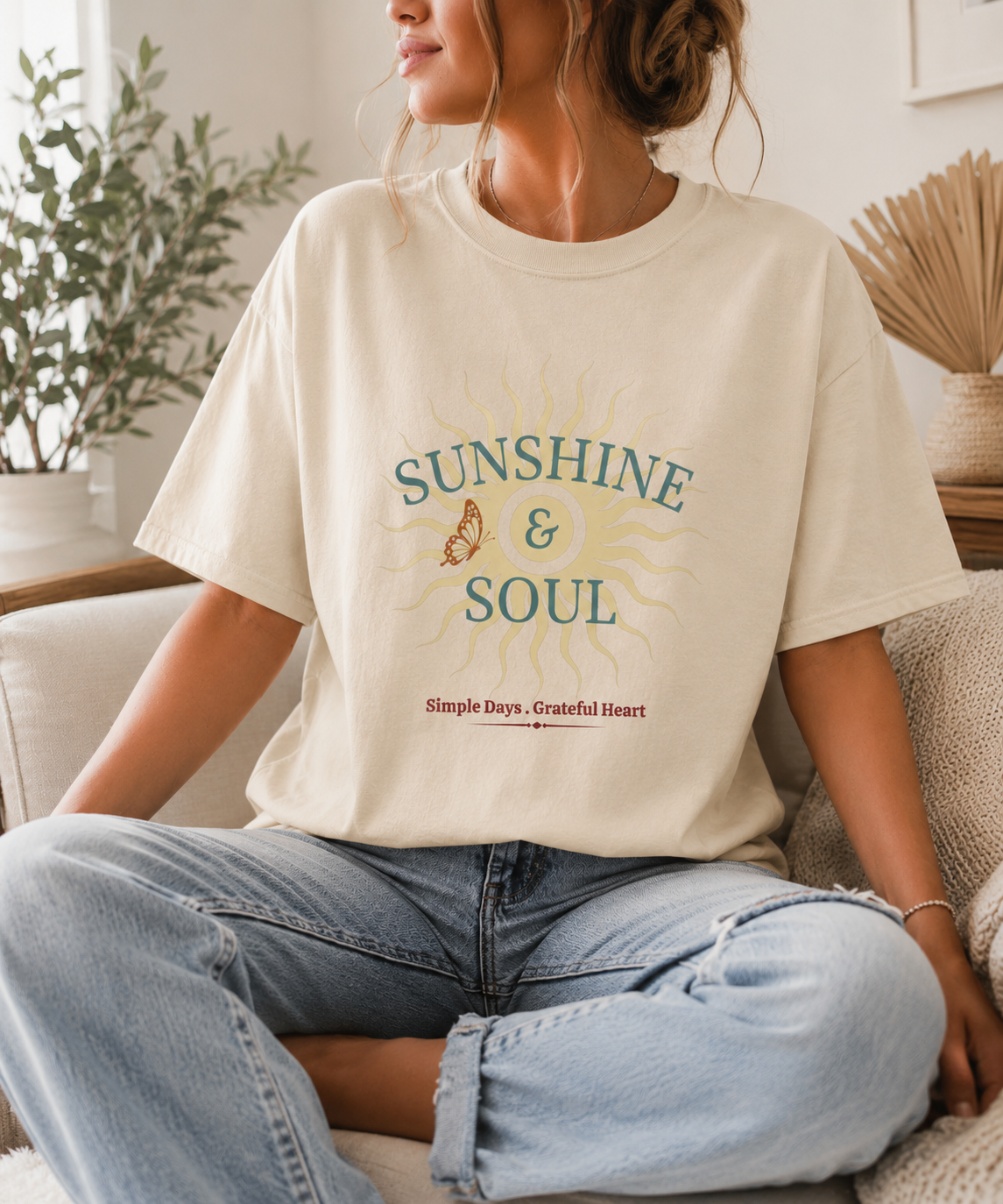 Lady on couch wearing a Sunshine and Soul Comfort Colors ivory t-shirt featuring vintage sun graphic with Simple Days Grateful Heart message in sage green and cream tones