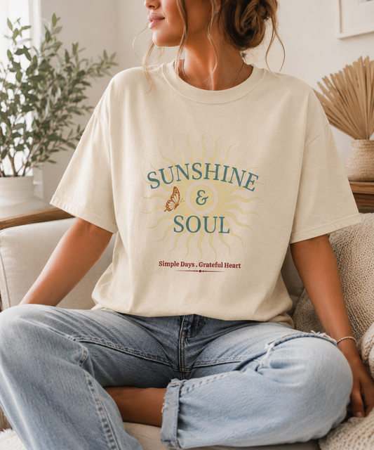 Lady on couch wearing a Sunshine and Soul Comfort Colors ivory t-shirt featuring vintage sun graphic with Simple Days Grateful Heart message in sage green and cream tones