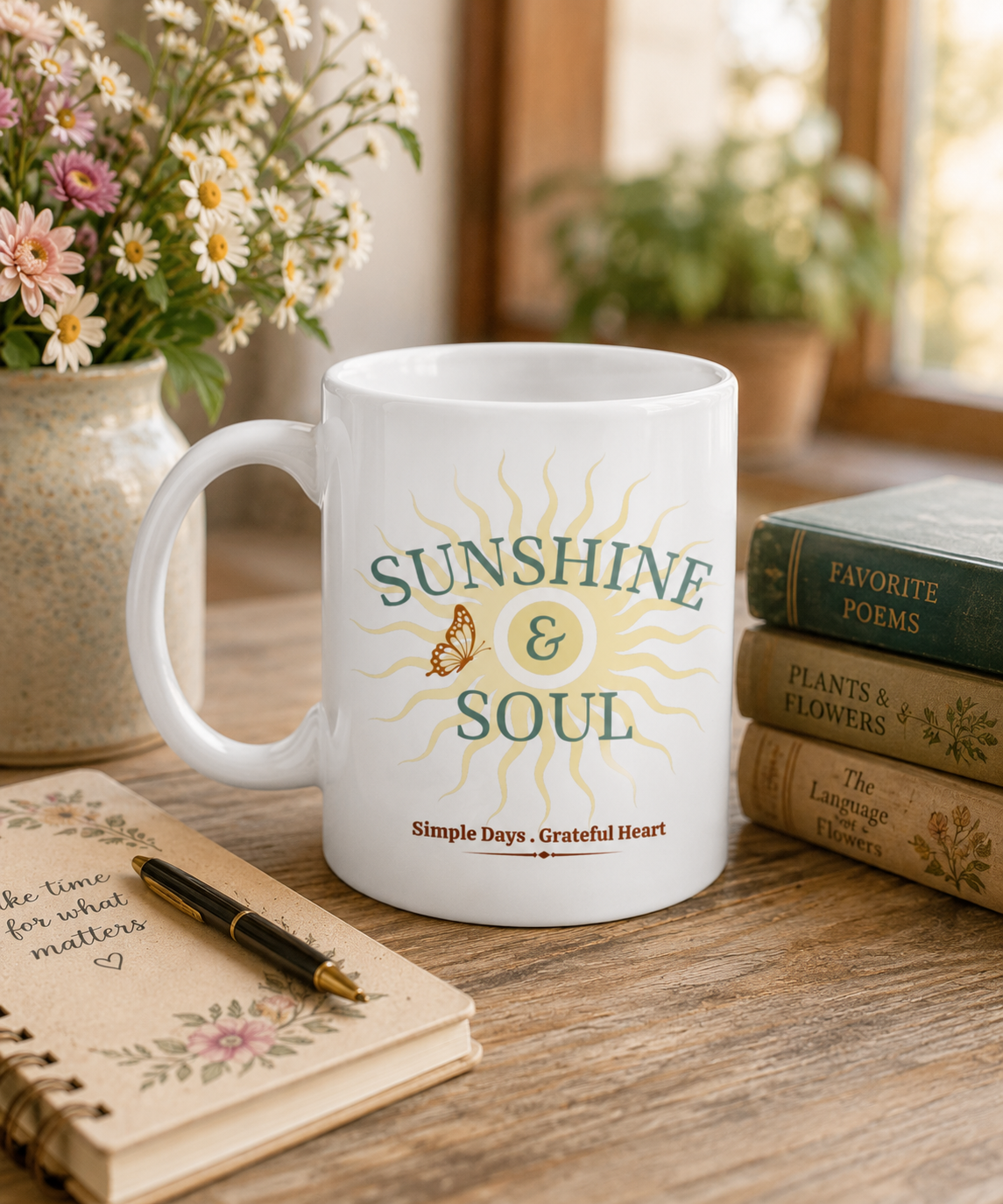 A white ceramic mug with a 'Sunshine & Soul' graphic sits on a wooden table surrounded by a notebook, pen, books, and a vase of flowers in a cozy, sunlit room.