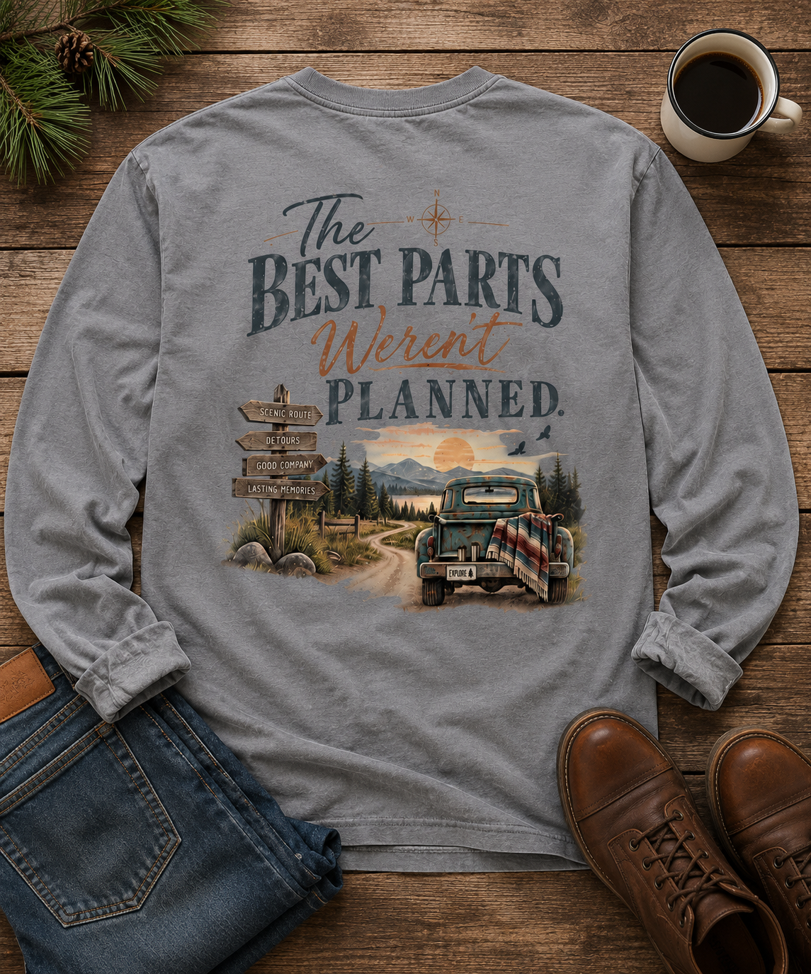 A flat lay of the back of a gray long sleeve t-shirt with a sunset mountain view and a dirt road . An old pickup truck and words that say The Best Parts Weren't Planned.